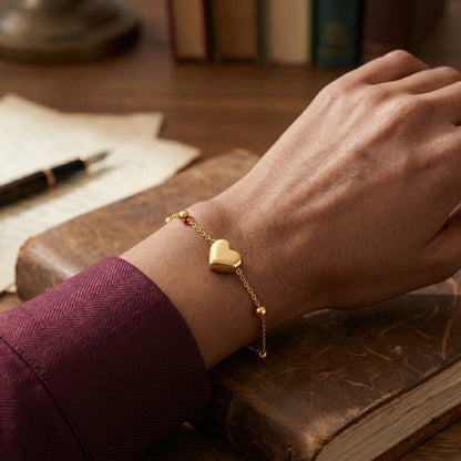 Hand wearing a gold heart-shaped bracelet on a wooden surface with books and a pen.