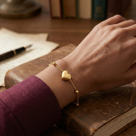 Hand wearing a gold heart-shaped bracelet on a wooden surface with books and a pen.